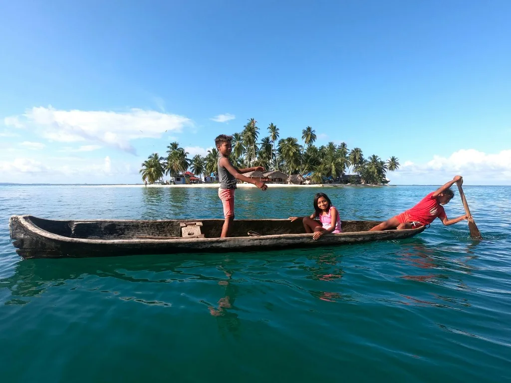 Three children in a wooden dugout canoe on turquoise water, with a palm island behind them